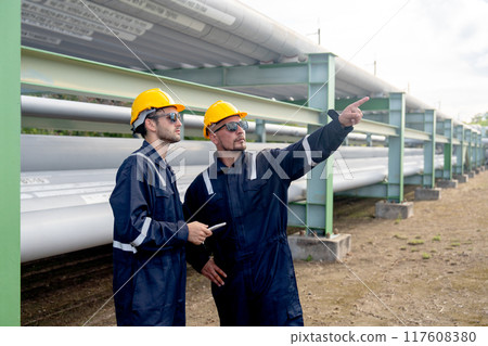 Two engineer or factory technician workers stand beside petrochemical gas pipeline and use tablet to discuss about their work with one point to right side. 117608380