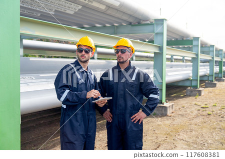 Two engineer or factory technician workers stand beside petrochemical gas pipeline and use tablet to discuss about their work. Two engineer or factory technician workers stand beside petrochemical gas pipeline and use tablet to discuss about their work. 117608381