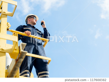 Factory engineer or technician worker man use walkie talkie to contact his team and stand on high state over pipeline of petrochemical gas in workplace with blue sky as background. Factory engineer or technician worker man use walkie talkie to contact his team and stand on high state over pipeline of petrochemical gas in workplace with blue sky as background. 117608382