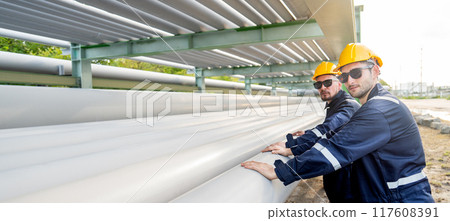 Side view of two engineer or factory technician workers stand beside petrochemical gas pipeline also touch and look at camera with warm light. 117608391