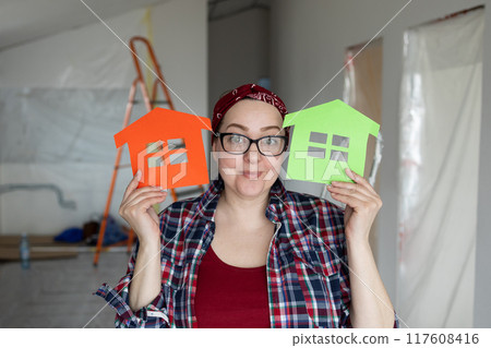 A Creative Woman Enthusiastically Holding Colorful House Cutouts in a Bright Renovation Space A Creative Woman Enthusiastically Holding Colorful House Cutouts in a Bright Renovation Space 117608416