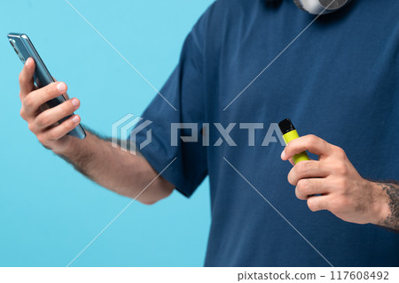 Green disposable electronic cigarette and phone in a man's hand. Modern online communication. on blue background Green disposable electronic cigarette and phone in a man's hand. Modern online communication. on blue background 117608492