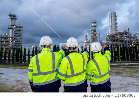 Back of engineer or technician workers stand with arm-crossed and one point to factory and look to petrochemical factory in the bakground with cloud sky. 117609003