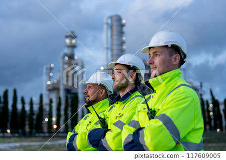 Close up factory engineer or technician workers stand in the row and look to left side with arm-crossed stand in front of refining and petrochemical factory during evening light. 117609005