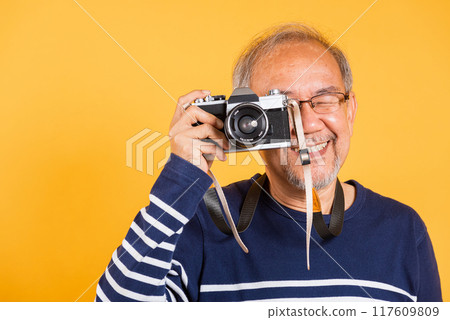 Portrait Asian old man wearing glasses look viewfinder vintage SLR camera studio shot isolated yellow background, smiling happy Photographer elderly man gray haired taking a picture of himself 117609809
