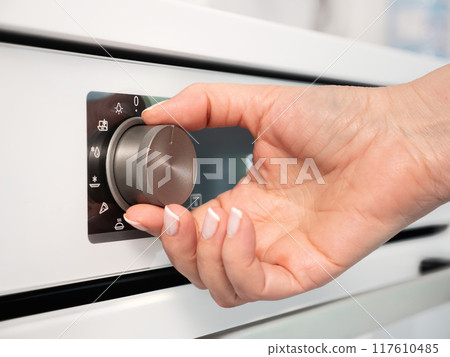 Woman adjusting modern white oven in kitchen.  Female hand pushing and turning knobs on oven control panel 117610485