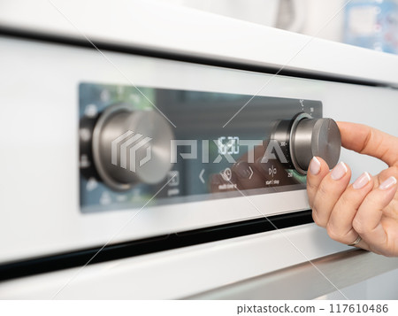 Woman adjusting modern white oven in kitchen.  Female hand pushing and turning knobs on oven control panel 117610486