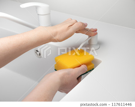 Female hand press on built-in dispenser in the sink with detergent. Woman washing dishes by yellow sponge with soap in white kitchen 117611446