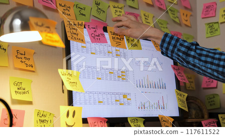 In a workspace filled with colorful papers, a young man attaches bright sticky notes to his computer screen 117611522