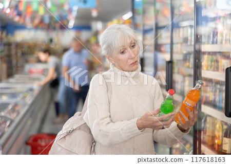 Old woman purchaser choosing cool drink in big supermarket Old woman purchaser choosing cool drink in big supermarket 117618163