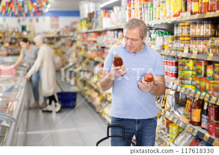Elderly man chooses and buys hot chili sauce in the grocery section of supermarket 117618330
