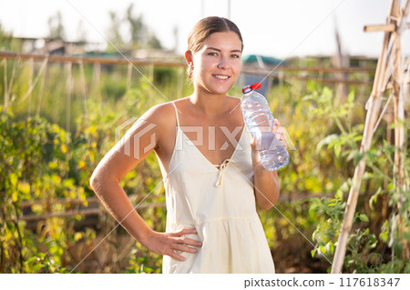 Hot summer weather in the garden beds of a country house - young woman drinks water from a bottle Hot summer weather in the garden beds of a country house - young woman drinks water from a bottle 117618347