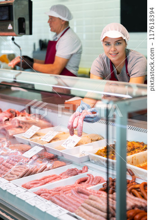 Female seller leans over to refrigerator window and takes out some raw barbecue sausages for buyer 117618394