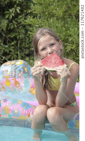 Schoolgirl in swimsuit eating watermelon near inflatable pool. Summer and vacation in yard of house. Lifestyle and rest.  117621482