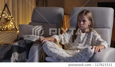 Schoolgirl watching movie in living room in pajamas in evening. Christmas and holidays. Modern interior and reclining rocking chair. Exciting pastime.  117621531