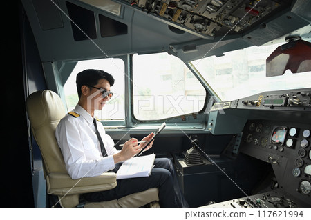 Handsome pilot wearing sunglasses using digital tablet, preparing for flight in the cockpit of an aircraft Handsome pilot wearing sunglasses using digital tablet, preparing for flight in the cockpit of an aircraft 117621994