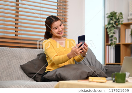 Happy young woman enjoying chatting with her friends using smartphone on sofa at home 117621999