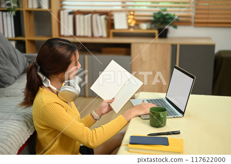 Focused female student holding notebook and working with laptop in living room 117622000