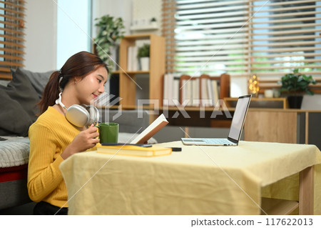Young woman with coffee cup sitting on the floor against a couch in living room and reading a book 117622013