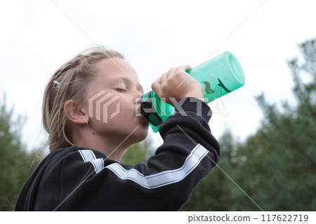 Schoolgirl in tracksuit drinking water from bottle. Sports in summer. Close-up portrait. Vacation and lifestyle.  117622719