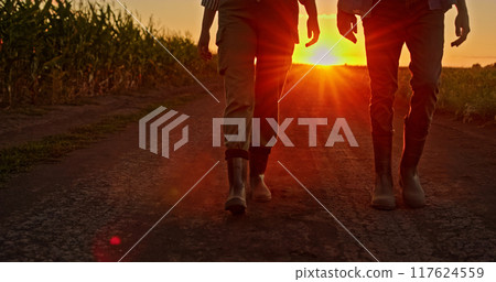 Legs of two farmers in boots walking on a dusty road through fields at sunset. Legs of two farmers in boots walking on a dusty road through fields at sunset. 117624559