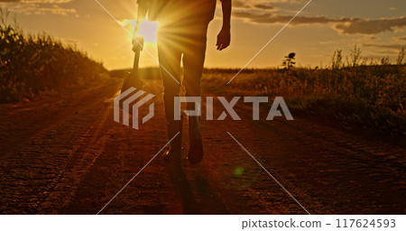Farmer's legs in boots walking through a field at sunset, carrying a shovel. 117624593
