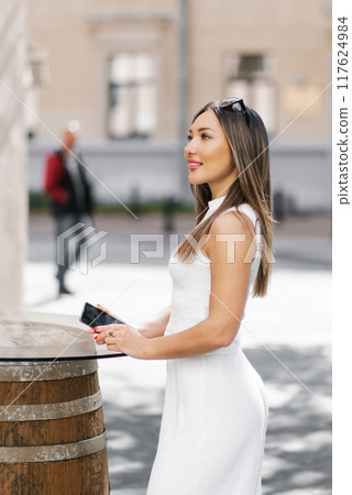 Smiling woman in casual clothes holds a mobile phone in her hands on a city street in summer, standing near a table of an outdoor cafe 117624984