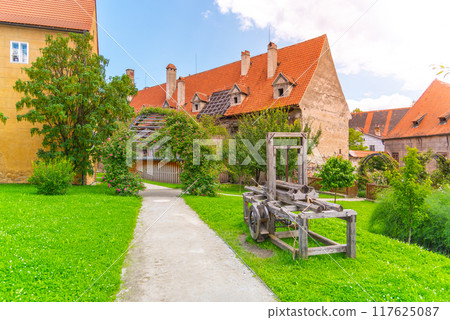 Visitors stroll through the lush grounds of a complex of three medieval monasteries in Cesky Krumlov, surrounded by historic buildings and greenery. A wooden exhibit adding to the rustic charm. Visitors stroll through the lush grounds of a complex of three medieval monasteries in Cesky Krumlov, surrounded by historic buildings and greenery. A wooden exhibit adding to the rustic charm. 117625087