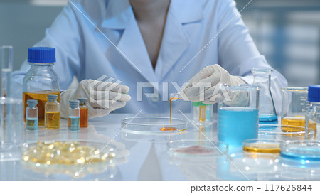 A scientist is pouring an orange solution into a petri dish for testing. Surrounding them are flasks containing test samples and substances. Creative pharmacy or health care concept. A scientist is pouring an orange solution into a petri dish for testing. Surrounding them are flasks containing test samples and substances. Creative pharmacy or health care concept. 117626844