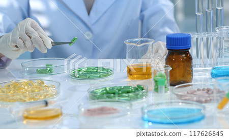 on the research table, many drug samples are stored in petri dishes. The researcher is using tweezers to pick up a green pill and examine it. Research landscape for medicinal products on the research table, many drug samples are stored in petri dishes. The researcher is using tweezers to pick up a green pill and examine it. Research landscape for medicinal products 117626845