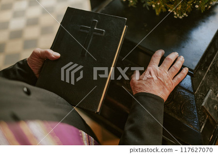 Unrecognizable priests wrinkled hand holding holy book and touching black coffin in church Unrecognizable priests wrinkled hand holding holy book and touching black coffin in church 117627070