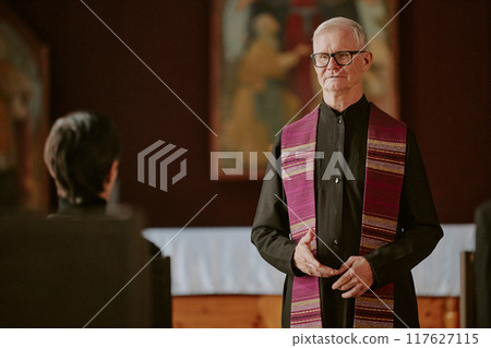 Silver-haired pastor dressed in black robe saying prayer during funeral 117627115