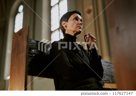 Senior woman dressed in total black sitting on wooden bench and holding wooden rosary in church Senior woman dressed in total black sitting on wooden bench and holding wooden rosary in church 117627147