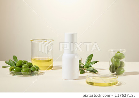 Front view, advertising photo, white cylindrical bottle placed in the middle on white table and white background, lab glassware containing fresh olives and olive oil decorated. 117627153