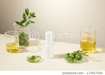 Cosmetic advertisement about olive ingredient, white cylindrical bottle placed in the middle of white table, surrounded by olive ingredients placed in glass laboratory equipment. 117627160