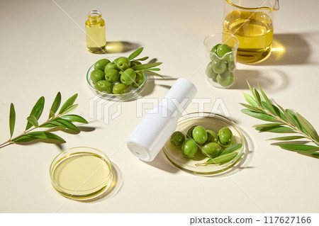 Top view, white cylindrical bottle is tilted onto a petri dish containing olives and essential oil, surrounded by olive branches and experimental glassware. Cosmetic natural theme. 117627166