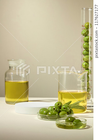 A tall graduated cylinder and petri dishes filled with green olives, beaker and glass jar filled with olive oil, empty white platform for products, ideal for cosmetics advertising. 117627177