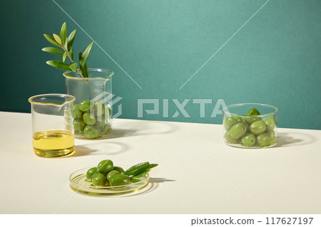 Fresh olives, oil, and branches are arranged in beakers and petri dishes on a white table, leaving room in the middle for product placement. Cosmetic advertisement Fresh olives, oil, and branches are arranged in beakers and petri dishes on a white table, leaving room in the middle for product placement. Cosmetic advertisement 117627197