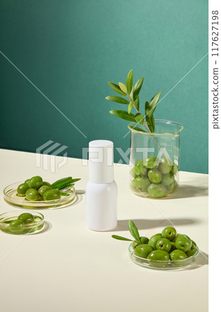 Simple cosmetic advertisement features a white cylindrical bottle, surrounded by petri dishes and beakers containing fresh green olives, olive leaves, and olive oil. Simple cosmetic advertisement features a white cylindrical bottle, surrounded by petri dishes and beakers containing fresh green olives, olive leaves, and olive oil. 117627198