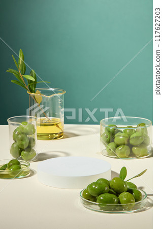 Minimalist advertising concept featuring fresh olives, branches, and oil on lab glassware, with white platform in middle, showcases products. 117627203