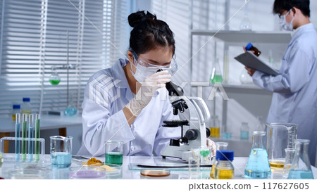 The scientist is looking at a microscope to study hair samples. Modern laboratory setting with chemicals and solutions to research hair care products, restore damaged hair The scientist is looking at a microscope to study hair samples. Modern laboratory setting with chemicals and solutions to research hair care products, restore damaged hair 117627605