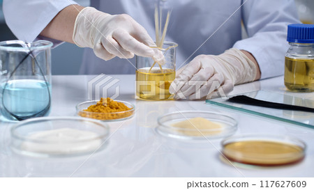 Front view of a scientist doing an experiment with a damaged hair sample. The scientist's hand is holding a sample of bleached hair dipped in a yellow solution Front view of a scientist doing an experiment with a damaged hair sample. The scientist's hand is holding a sample of bleached hair dipped in a yellow solution 117627609