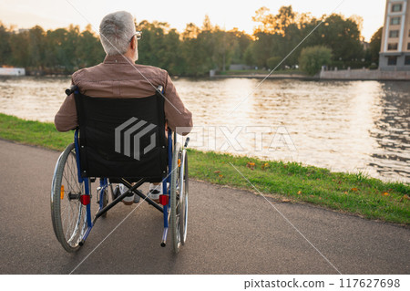 Sad senior man sitting in wheelchair walking on road in city park outdoor, back view. Old paralyzed man in chair for people with disability feel depressed. Lonely elderly sick man sits on wheelchair 117627698