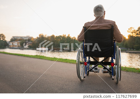 Sad senior man sitting in wheelchair walking on road in city park outdoor, back view. Old paralyzed man in chair for people with disability feel depressed. Lonely elderly sick man sits on wheelchair 117627699