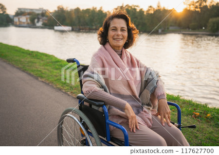 Happy senior woman in wheelchair smiling enjoying freedom walking on road in city park outdoor. Old cheerful positive woman in chair for people with disability. Disability disease rehabilitation Happy senior woman in wheelchair smiling enjoying freedom walking on road in city park outdoor. Old cheerful positive woman in chair for people with disability. Disability disease rehabilitation 117627700