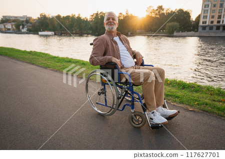 Happy senior man in wheelchair smiling enjoying freedom walking on road in city park outdoor. Old cheerful positive man in chair for people with disability. Disability disease rehabilitation Happy senior man in wheelchair smiling enjoying freedom walking on road in city park outdoor. Old cheerful positive man in chair for people with disability. Disability disease rehabilitation 117627701
