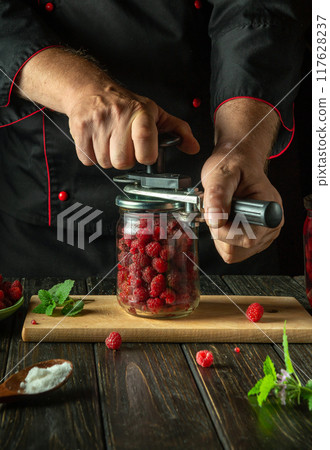 A chef rolls up a jar of ripe raspberries for canning with a hand seaming key. Making or canning raspberry jam or compote in the kitchen 117628237