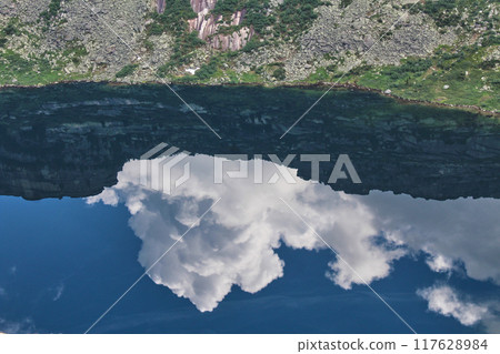 Bright mirror reflection of white clouds and blue sky in water of a mountain lake. Summer landscape in nature park Bright mirror reflection of white clouds and blue sky in water of a mountain lake. Summer landscape in nature park 117628984