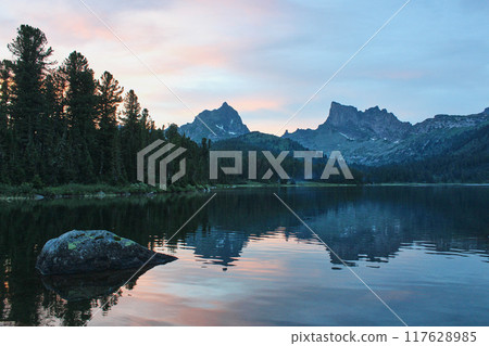 Two rocky mountain peaks are reflected in the water of the lake on summer sunset 117628985