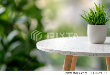 A small potted plant on a light table surrounded by green foliage indoors 117629163